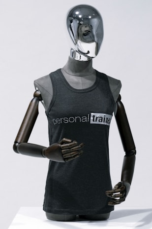 A metallic mannequin with a smooth, reflective head and dark, articulated arms is wearing a sleeveless shirt that reads 'personal trainer'. The mannequin stands upright against a plain light background, emphasizing the sleek design.