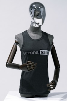 A metallic mannequin with a smooth, reflective head and dark, articulated arms is wearing a sleeveless shirt that reads 'personal trainer'. The mannequin stands upright against a plain light background, emphasizing the sleek design.