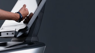 A person interacting with a touchscreen monitor, possibly adjusting its angle. The image focuses on the hand wearing a smartwatch, with the dark background emphasizing the device's modern design.