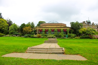 A well-maintained garden with lush green grass and a variety of vibrant plants and flowers, leading to a round, elegant building with a domed roof and large windows. Stone steps and a pathway are in the foreground, surrounded by trees and bushes.