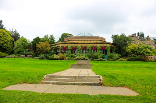 A well-maintained garden with lush green grass and a variety of vibrant plants and flowers, leading to a round, elegant building with a domed roof and large windows. Stone steps and a pathway are in the foreground, surrounded by trees and bushes.
