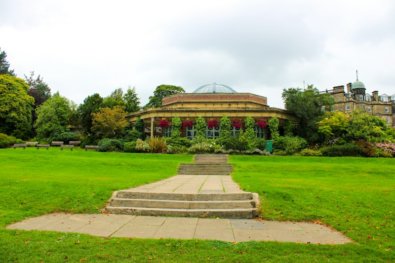 A well-maintained garden with lush green grass and a variety of vibrant plants and flowers, leading to a round, elegant building with a domed roof and large windows. Stone steps and a pathway are in the foreground, surrounded by trees and bushes.