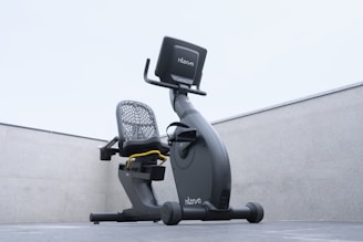 A modern recumbent exercise bike is positioned outdoors on a concrete surface. The bike has a sleek design with a mesh backrest and an electronic display screen. The environment is minimalist, with the bike framed by two concrete walls and an overcast sky above.