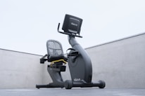 A modern recumbent exercise bike is positioned outdoors on a concrete surface. The bike has a sleek design with a mesh backrest and an electronic display screen. The environment is minimalist, with the bike framed by two concrete walls and an overcast sky above.