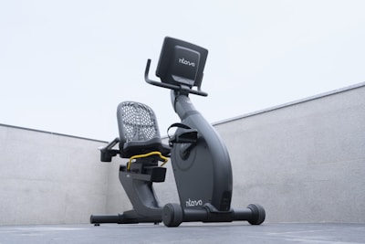 A modern recumbent exercise bike is positioned outdoors on a concrete surface. The bike has a sleek design with a mesh backrest and an electronic display screen. The environment is minimalist, with the bike framed by two concrete walls and an overcast sky above.