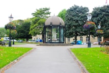 A well-maintained urban park with green lawns, trees, and benches. A central feature is a glass pavilion containing a statue. Surrounding the pavilion are flower-filled street lamps and several parked vehicles can be seen in the background near a building.