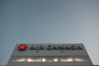 A large building facade with the Air Canada logo prominently displayed. The logo features a red maple leaf within a circle. The building is constructed from reflective materials and the scene is captured under a clear sky, suggesting a modern and corporate environment.