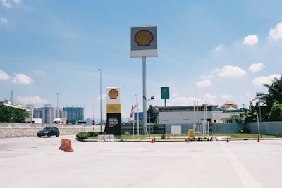 A bright and clear day at a Shell gas station, with a large sign displaying the Shell logo prominently. Several flagpoles with flags are in view, along with a Petronas sign in the background. Some construction is visible beyond the compound, and there is a car driving past on the road.