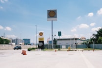 A bright and clear day at a Shell gas station, with a large sign displaying the Shell logo prominently. Several flagpoles with flags are in view, along with a Petronas sign in the background. Some construction is visible beyond the compound, and there is a car driving past on the road.