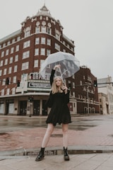 A person stands confidently on a wet city street holding a transparent umbrella. They are wearing a black dress and boots. Behind them is a tall, historic building made of red brick with ornate architectural details. The ground is reflective from the rain, and the sky is overcast.