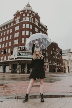 A person stands confidently on a wet city street holding a transparent umbrella. They are wearing a black dress and boots. Behind them is a tall, historic building made of red brick with ornate architectural details. The ground is reflective from the rain, and the sky is overcast.