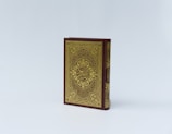 An ornate book with a richly decorated cover, featuring intricate gold patterns and Arabic calligraphy on a dark maroon background. The book is positioned upright against a plain, light background.