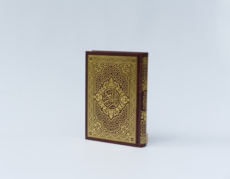 An ornate book with a richly decorated cover, featuring intricate gold patterns and Arabic calligraphy on a dark maroon background. The book is positioned upright against a plain, light background.