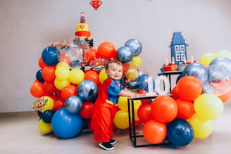 A child dressed in a superhero costume stands surrounded by colorful balloons and decorations featuring comic book elements. The background includes a cake with superhero motifs and a decorative stand with cupcakes.