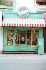 A storefront with a colorful and retro design. The sign above reads 'Treats & Sweets' and features a candy emblem. The store has a bright turquoise exterior with yellow trimming and a red-and-white striped awning. Large windows display various colorful signs for candy and sweets. A small potted plant sits to the right of the entrance.