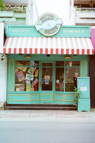 A storefront with a colorful and retro design. The sign above reads 'Treats & Sweets' and features a candy emblem. The store has a bright turquoise exterior with yellow trimming and a red-and-white striped awning. Large windows display various colorful signs for candy and sweets. A small potted plant sits to the right of the entrance.