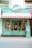 A storefront with a colorful and retro design. The sign above reads 'Treats & Sweets' and features a candy emblem. The store has a bright turquoise exterior with yellow trimming and a red-and-white striped awning. Large windows display various colorful signs for candy and sweets. A small potted plant sits to the right of the entrance.