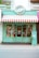A storefront with a colorful and retro design. The sign above reads 'Treats & Sweets' and features a candy emblem. The store has a bright turquoise exterior with yellow trimming and a red-and-white striped awning. Large windows display various colorful signs for candy and sweets. A small potted plant sits to the right of the entrance.