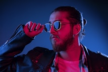 A person with a beard wearing a leather jacket and sunglasses is posing. The lighting casts a dramatic red and blue color contrast, creating a vibrant atmosphere. The person's hair is tied back and they are adjusting their sunglasses with one hand, imparting a cool and confident look.