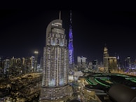 A nighttime cityscape featuring modern skyscrapers illuminated with lights, including a prominent tall tower with 'EMAAR' written on it. The skyline is dotted with various buildings, and the foreground shows a mix of architectural styles.