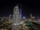 A nighttime cityscape featuring modern skyscrapers illuminated with lights, including a prominent tall tower with 'EMAAR' written on it. The skyline is dotted with various buildings, and the foreground shows a mix of architectural styles.
