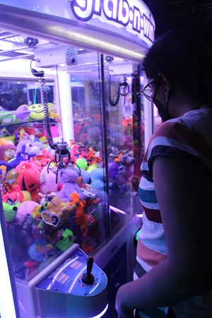 A person is standing in front of a claw machine filled with colorful stuffed toys. The machine is lit with bright purple and blue lights, and the individual is holding the joystick controller. The toys inside the machine include various shapes, sizes, and colors, contributing to a vibrant scene.