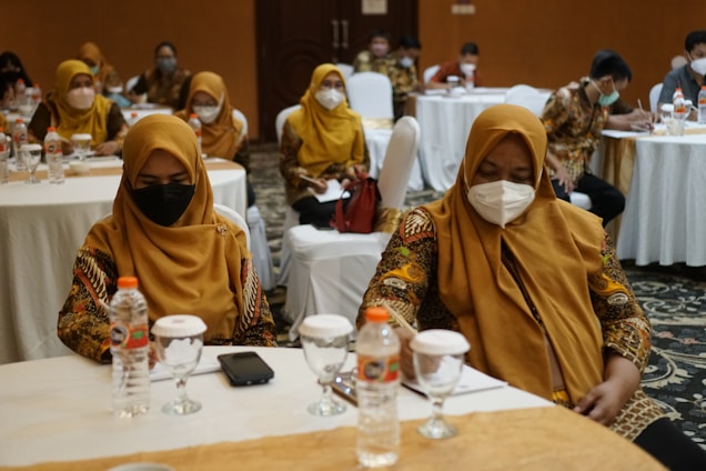 A group of people is seated at round tables in a formal setting, with most wearing patterned attire and face masks. The room contains white chairs and tables, with water bottles and notepads present. A conference or meeting appears to be taking place.
