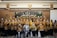 A group of people is gathered for a sensitivity training session, posing together for a photo. Many are wearing uniform-like attire in brown and yellow, and most are wearing face masks. They appear to be in a conference room with a banner behind them and portraits hanging on the wall.