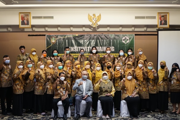 A group of people is gathered for a sensitivity training session, posing together for a photo. Many are wearing uniform-like attire in brown and yellow, and most are wearing face masks. They appear to be in a conference room with a banner behind them and portraits hanging on the wall.