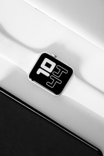 A modern digital smartwatch with a white strap is displayed from a close-up angle. The watch face prominently shows the time with bold, white digital numbers on a black background. The top and bottom of the image have a mix of white and black surfaces, creating a minimalist and sleek aesthetic.