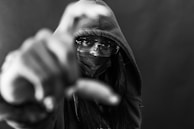 A person wearing a hooded sweatshirt and sunglasses appears to be reaching towards the camera with a hand in the foreground. Their face is partially covered by a mask, creating a sense of mystery.