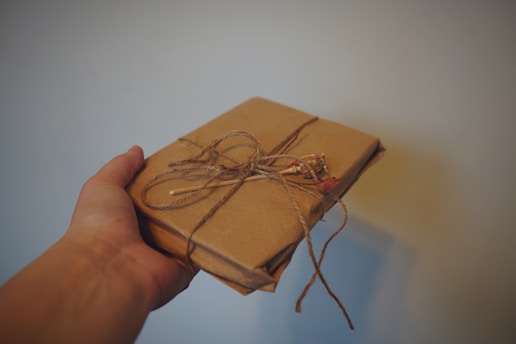 A hand is holding a small package wrapped in brown paper and secured with twine. The package is simply decorated with a twig and a small sprig of dried flowers.