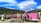 A vibrant outdoor display features numerous bras hung on a wire fence against a scenic backdrop of rolling green hills and a striking blue sky with unique cloud formations. A pink sign reads 'Welcome to Bradrona' and encourages donations for breast cancer support. A small sculptural figure sits on the left.