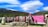 A vibrant outdoor display features numerous bras hung on a wire fence against a scenic backdrop of rolling green hills and a striking blue sky with unique cloud formations. A pink sign reads 'Welcome to Bradrona' and encourages donations for breast cancer support. A small sculptural figure sits on the left.