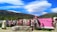 A vibrant outdoor display features numerous bras hung on a wire fence against a scenic backdrop of rolling green hills and a striking blue sky with unique cloud formations. A pink sign reads 'Welcome to Bradrona' and encourages donations for breast cancer support. A small sculptural figure sits on the left.