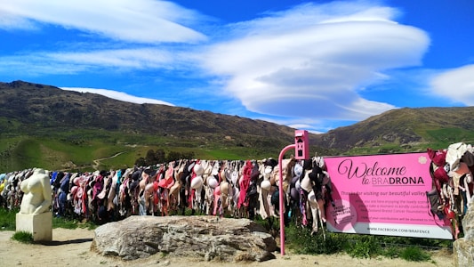 A vibrant outdoor display features numerous bras hung on a wire fence against a scenic backdrop of rolling green hills and a striking blue sky with unique cloud formations. A pink sign reads 'Welcome to Bradrona' and encourages donations for breast cancer support. A small sculptural figure sits on the left.