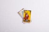 A set of tarot cards spread in a fan-like arrangement with the card labeled 'The Magician' prominently displayed on top. The card features an illustration of a figure in red robes with one hand raised, holding a wand, surrounded by various symbolic items. The background is predominantly yellow with decorative elements.