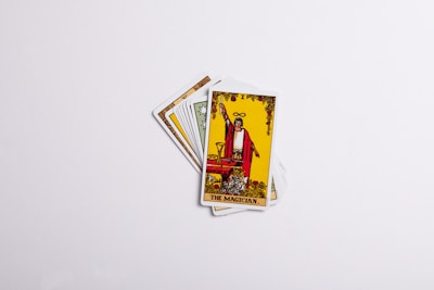A set of tarot cards spread in a fan-like arrangement with the card labeled 'The Magician' prominently displayed on top. The card features an illustration of a figure in red robes with one hand raised, holding a wand, surrounded by various symbolic items. The background is predominantly yellow with decorative elements.