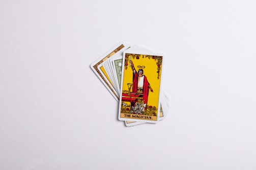 A set of tarot cards spread in a fan-like arrangement with the card labeled 'The Magician' prominently displayed on top. The card features an illustration of a figure in red robes with one hand raised, holding a wand, surrounded by various symbolic items. The background is predominantly yellow with decorative elements.