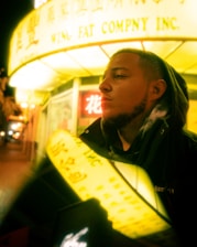 A person with a contemplative expression stands outside a building with bright yellow signage displaying both Chinese and English text. The illumination from the signs casts a warm glow over the scene, adding an urban and lively atmosphere.
