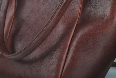 A close-up view of a leather surface with a rich, brown texture. The material appears smooth with visible grain patterns, highlighting the quality of the leather. The image captures part of a strap, suggesting it's a piece of leather goods, possibly a bag.