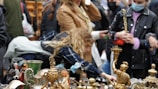 People gather around a stall with various vintage and antique items on display, including decorative brass figurines and pottery. Some individuals are closely inspecting the items, suggesting a lively market atmosphere.