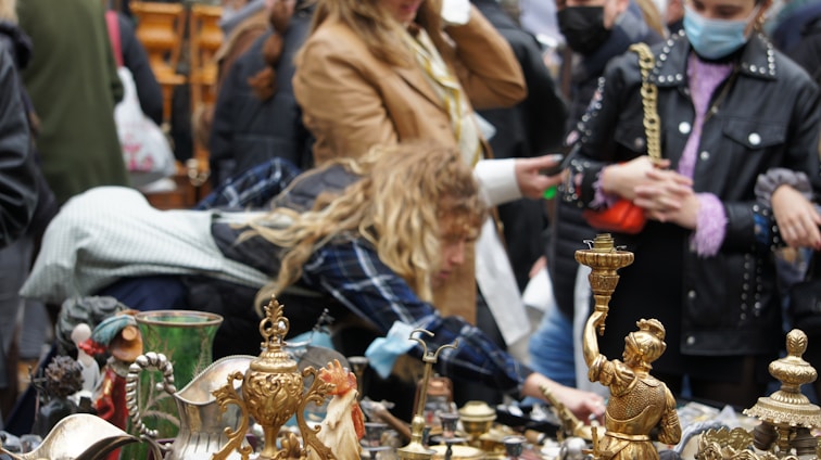 People gather around a stall with various vintage and antique items on display, including decorative brass figurines and pottery. Some individuals are closely inspecting the items, suggesting a lively market atmosphere.