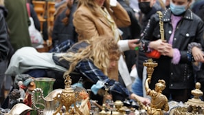 People gather around a stall with various vintage and antique items on display, including decorative brass figurines and pottery. Some individuals are closely inspecting the items, suggesting a lively market atmosphere.