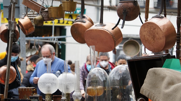 A bustling street market scene with several copper pots and pans hanging prominently in the foreground. There are also vintage glass lamps and various metal objects displayed. People in masks are observed in the background, indicating a crowded and lively atmosphere.