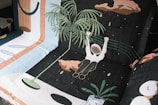 A woven fabric features a whimsical scene with an astronaut-like figure floating among abstract shapes and designs. The astronaut is holding onto a leafy plant, surrounded by pastel and earth-toned geometric patterns on a dark background.
