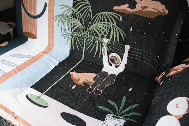 A woven fabric features a whimsical scene with an astronaut-like figure floating among abstract shapes and designs. The astronaut is holding onto a leafy plant, surrounded by pastel and earth-toned geometric patterns on a dark background.