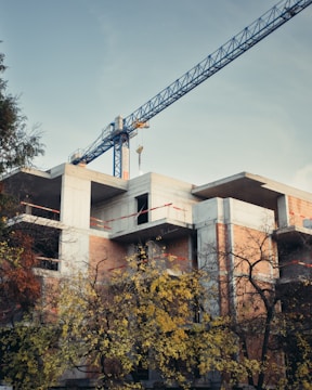 A construction site with a partially built concrete structure surrounded by trees. A large blue crane is positioned over the building, indicating ongoing construction work. The lower part of the building is obscured by trees with some yellow and brown foliage.