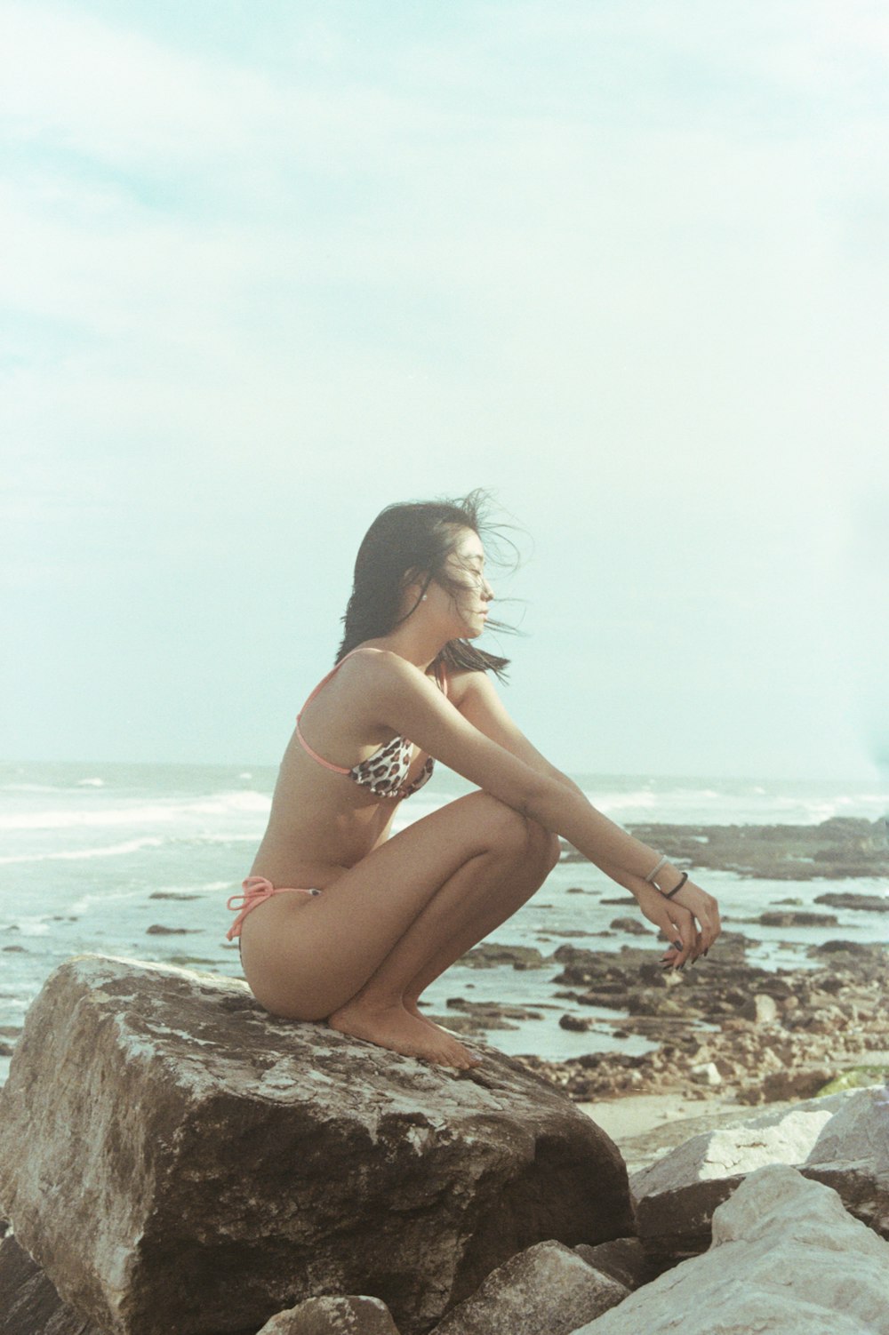 a woman in a bikini sitting on a rock by the ocean