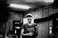 A black and white photograph of a person smiling broadly, holding a camera. The setting appears to be indoors with brick walls and a dimly lit atmosphere. The person is wearing a cap and a sleeveless shirt with visible tattoos on their arms. There are shelves and other objects in the background.